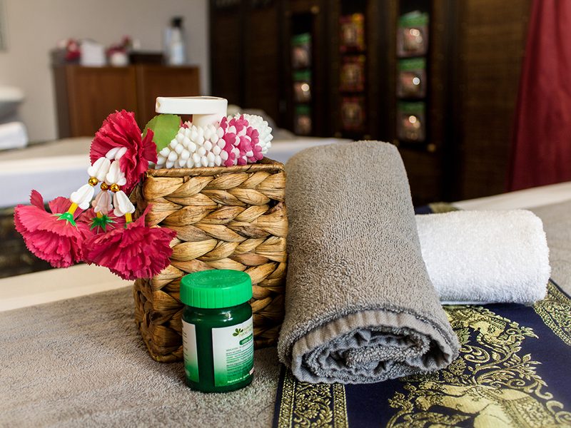 Korb mit Massageölen, Blütenkette und gerollten Handtüchern.